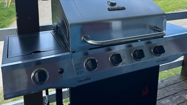 A Char-Broil gas grill with four burners, a side burner, and a chimney, set on a wooden deck with a view of a house and water body in the background. - Colonels Quarters of Eastport (Eastport)