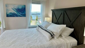 A neatly made bed with white linens and a striped pillow, a dark wooden headboard, a painting of a wave on the wall, and a window with curtains. - Colonels Quarters of Eastport (Eastport)