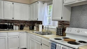 A kitchen with white cabinets, a stove, a sink, and a window with curtains. - Colonels Quarters of Eastport (Eastport)