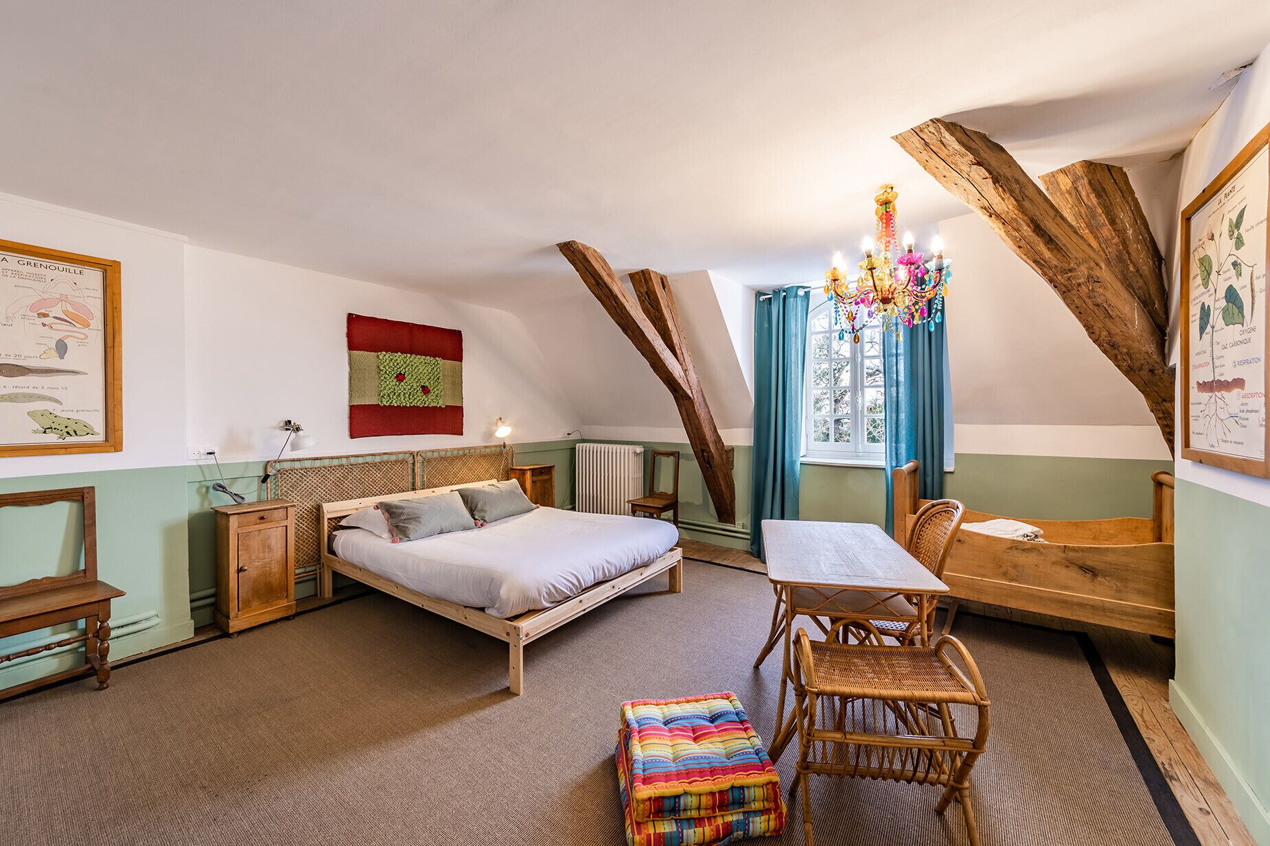 A cozy bedroom with a bed, a small table, and a chair. There is a window with curtains, a radiator, and a wooden beam structure.