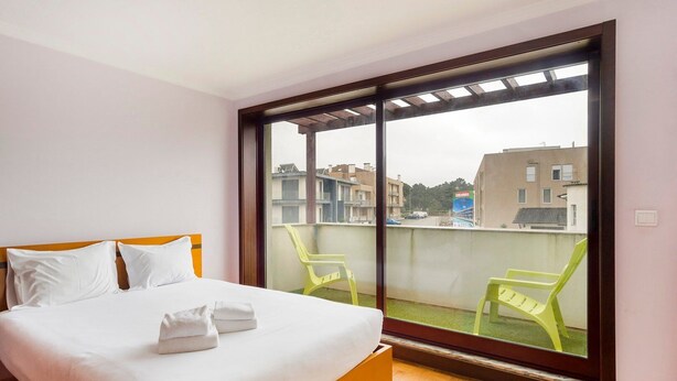 A hotel room with a bed, a balcony with chairs, and a view of buildings. - GuestReady - Comfortable Retreat in Esposende (Esposende)