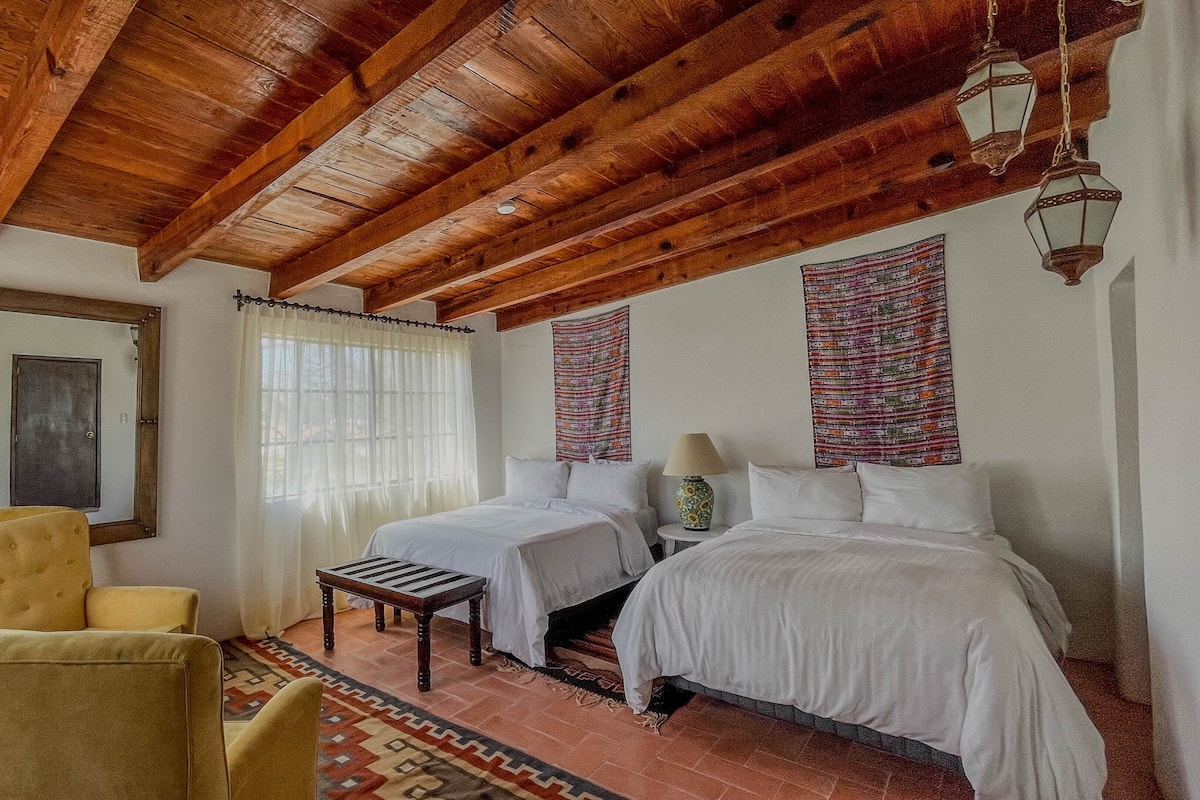 A room with a wooden ceiling, two beds, a yellow armchair, a small table, and a patterned rug.
