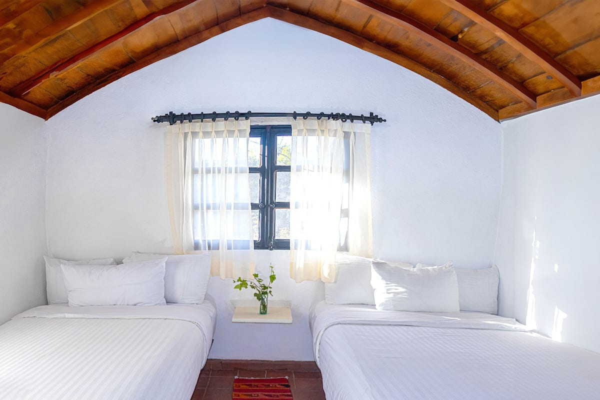 A room with two beds, a window with curtains, and a wooden ceiling.
