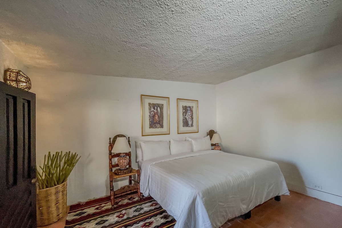 A bedroom with a bed, two framed pictures, a nightstand, a lamp, a wicker basket, and a patterned rug.