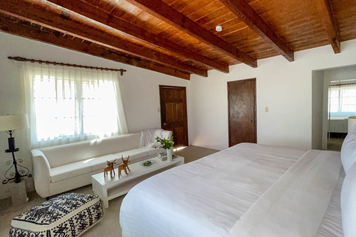 A bedroom with a large bed, a sofa, a small table with decorative items, and a window with curtains.