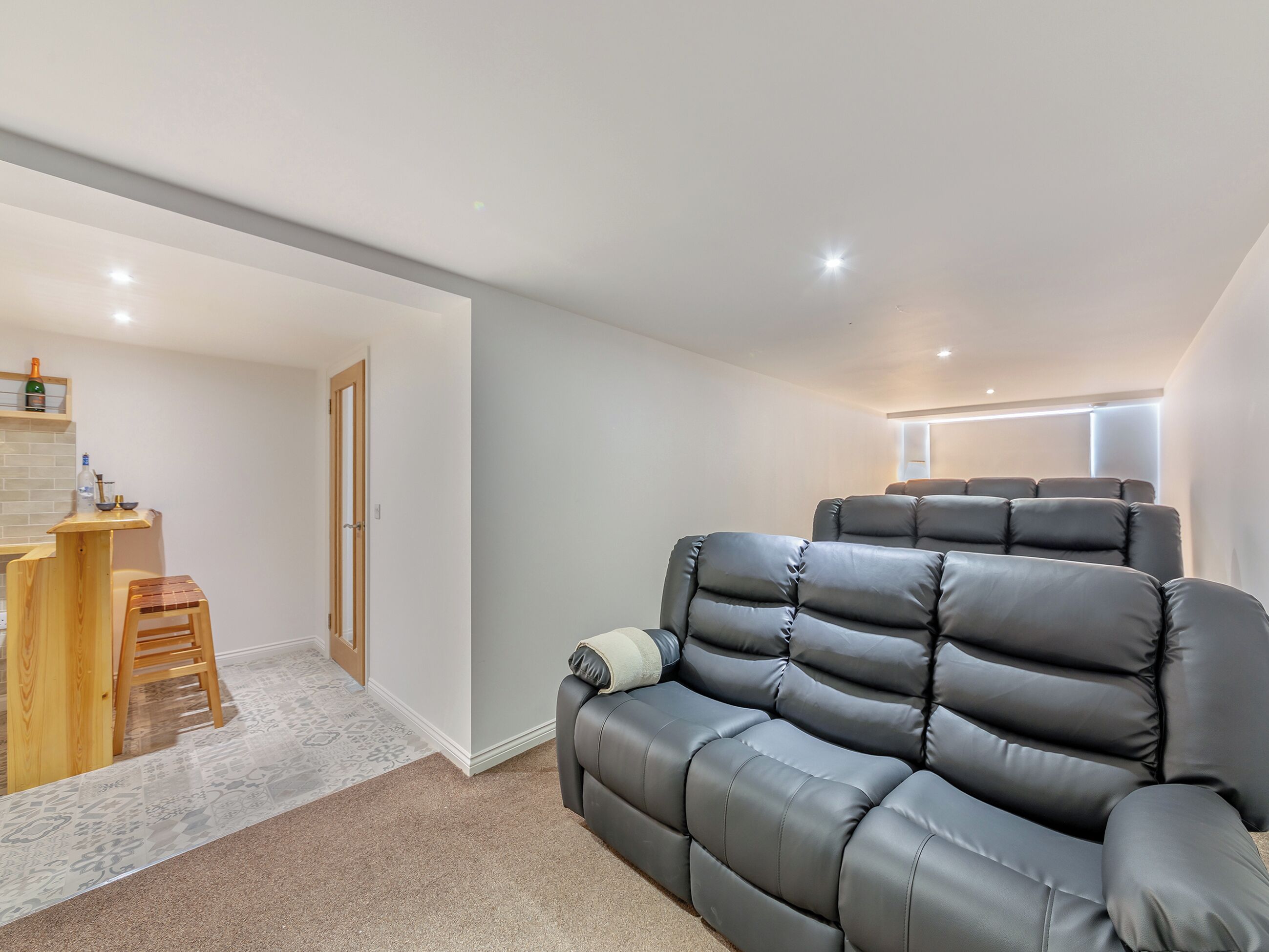 A modern living room with a grey leather sofa, a wooden coffee table, and a small kitchen area with a shelf.