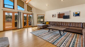 A spacious living room with a brown leather sofa, a wooden coffee table, and a patterned rug. Large windows and a sliding glass door provide natural light and views of the outdoors.