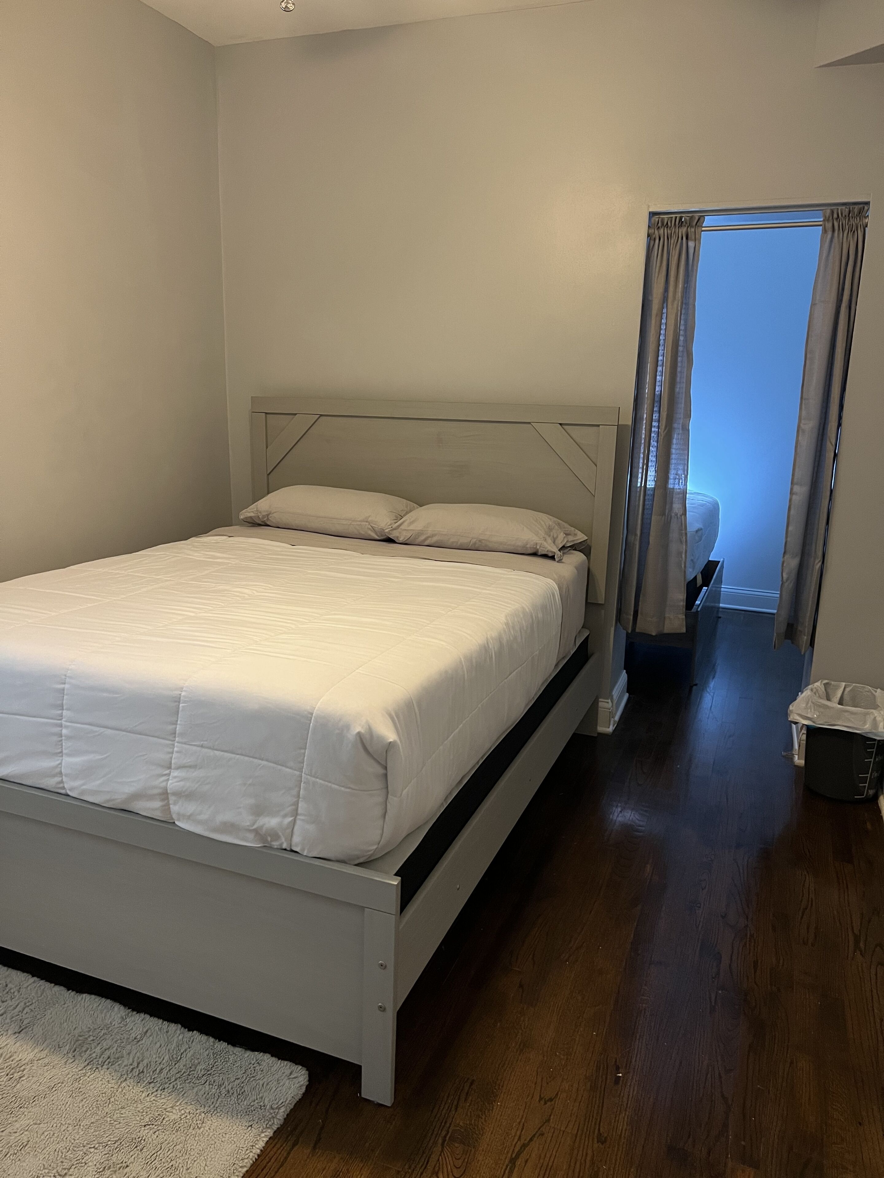 Een slaapkamer met een bed, een wit hoofdbord, een houten vloer en uitzicht naar buiten door een raam met gordijnen.