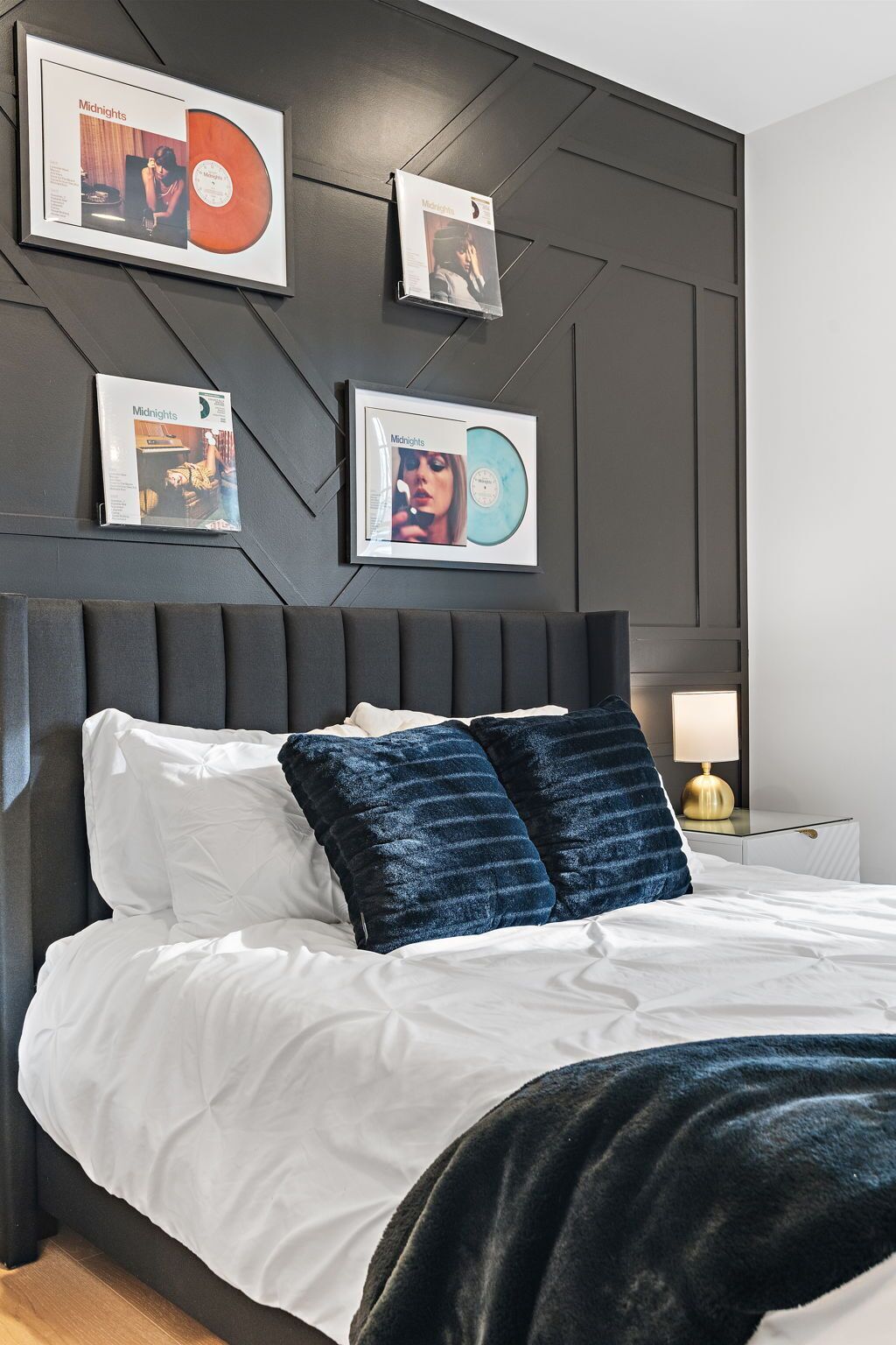A bedroom with a bed, pillows, a nightstand, a lamp, and framed records on the wall.