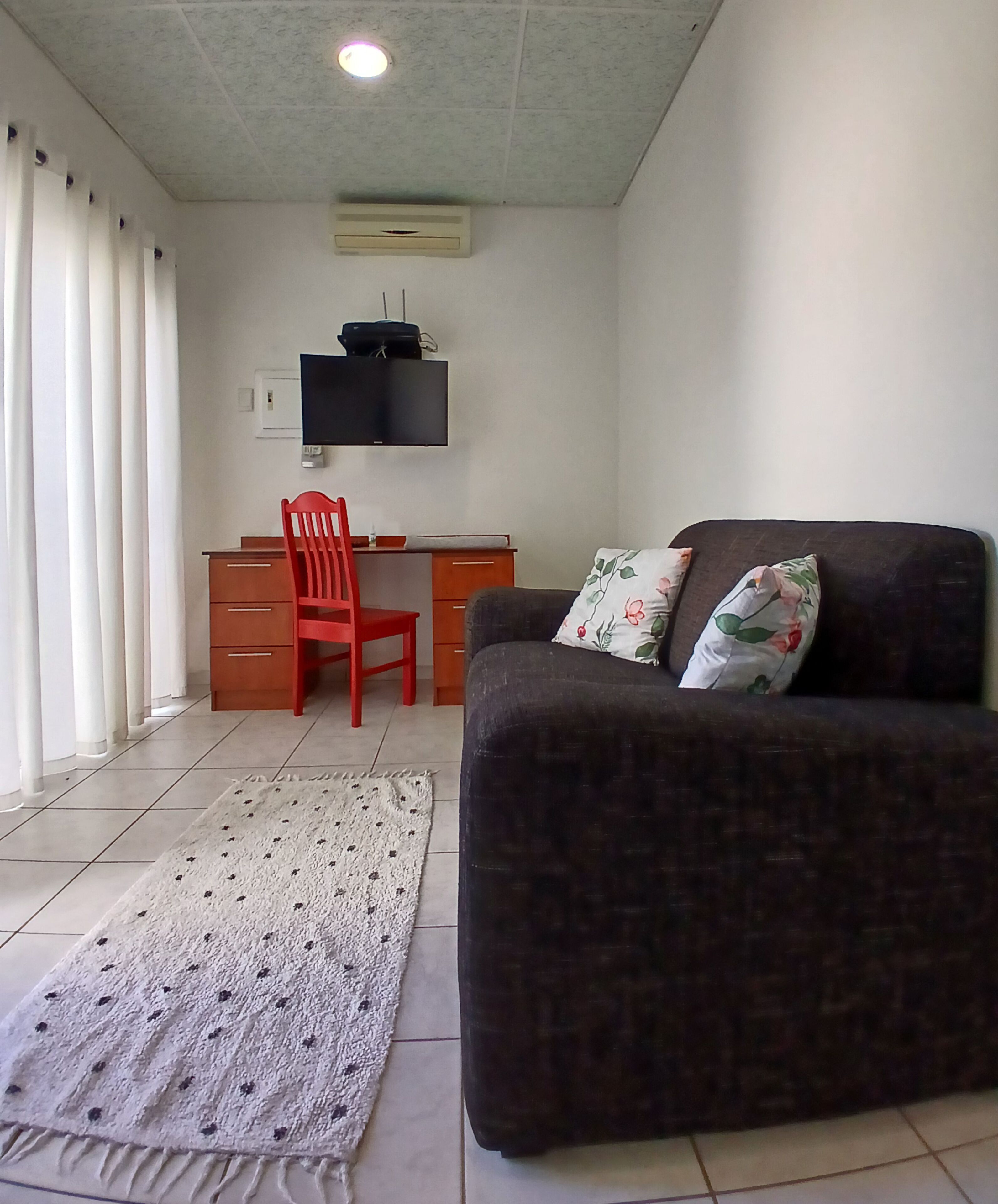 Una habitación con un sofá marrón oscuro, una silla roja, un escritorio y un televisor de pantalla plana montado en la pared.