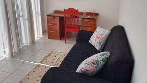 Una habitación con un sofá negro, un escritorio de madera, una silla roja, un televisor de pantalla plana y un ventanal con cortinas.