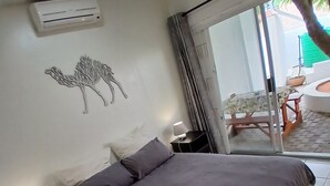 Un dormitorio con cama, mesita de noche, lámpara, aire acondicionado instalado en la pared y ventana con vistas al exterior.
