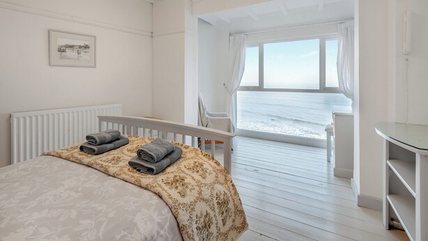 Un dormitorio con cama, vista al mar, una ventana con cortinas y una pequeña mesa.