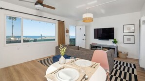 A modern living room with a dining area, a television, and a view of the beach. - 706 S Pacific Unit C · Steps From Beach, Panoramic Ocean Views & Parking (Oceanside)