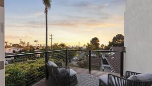 Ein Balkon mit Korbmöbeln, einem gemusterten Teppich und Blick auf eine Wohngegend bei Sonnenuntergang.