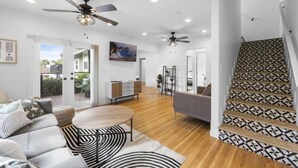 Ein geräumiger Wohnbereich mit Sofa, Couchtisch und einem gemusterten Teppich. Deckenventilatoren, ein Fernseher und eine Treppe mit gemusterten Fliesen sind vorhanden.
