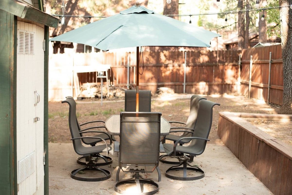 An outdoor patio with a table and four chairs, a blue umbrella, and a wooden fence in the background.