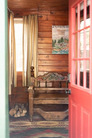 A wooden cabin interior with a red door, a bench, a poster, and a window with curtains.