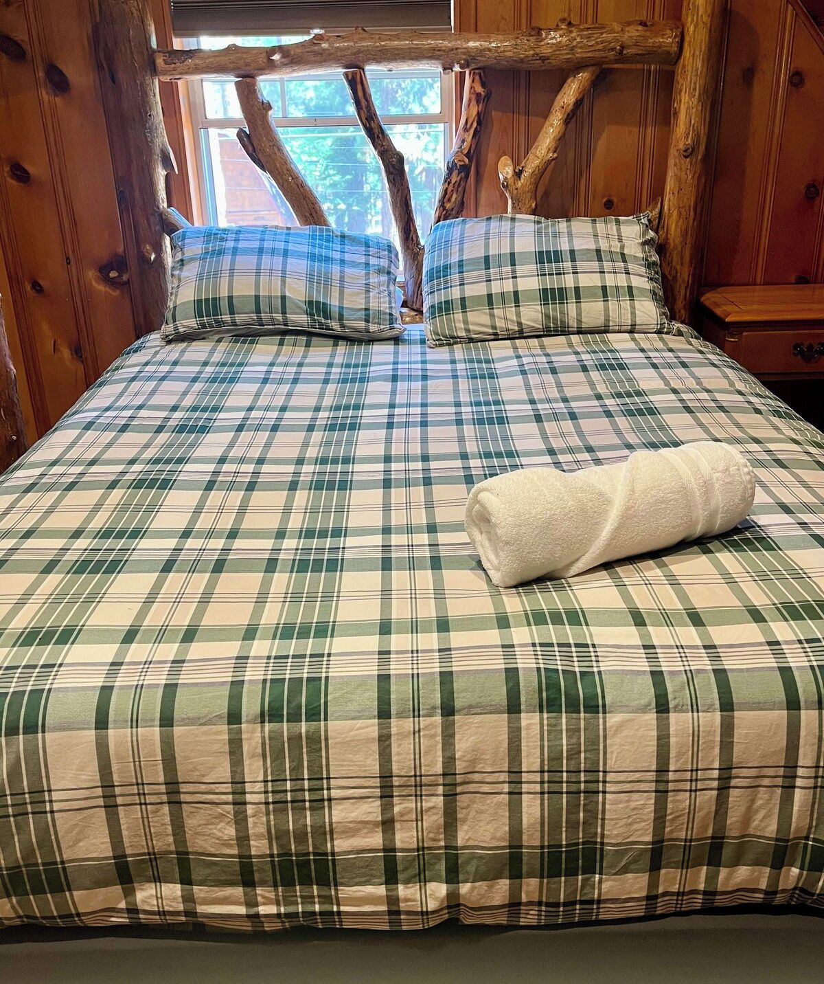 A bed with a plaid bedspread and pillows, a wooden headboard, and a window with a view of trees.