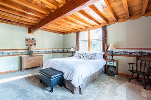 A bedroom with a wooden ceiling, a bed with white bedding, a nightstand, a lamp, and a window with a view.