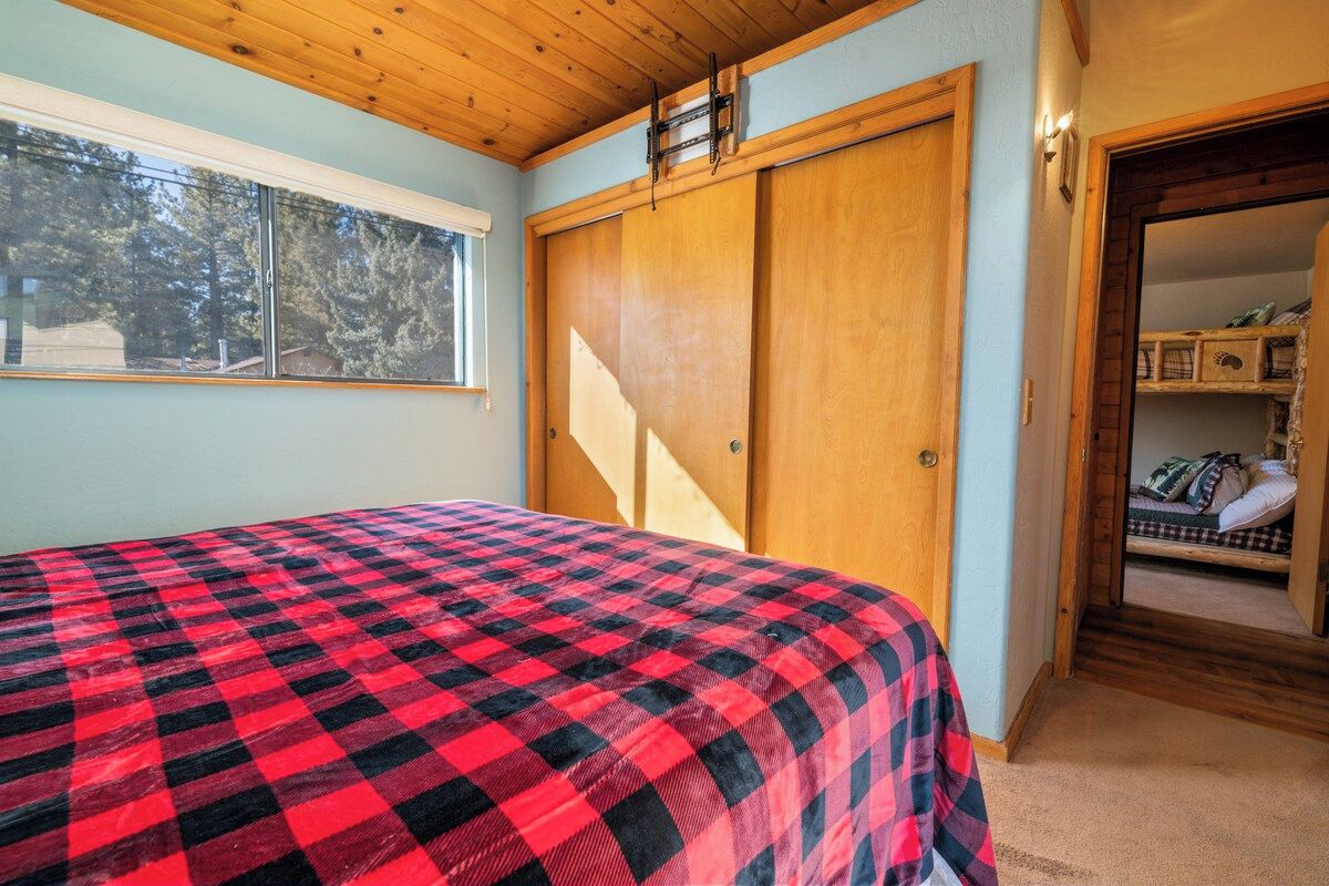 A bedroom with a checkered bedspread, a wooden wardrobe, and a window with a view of trees.