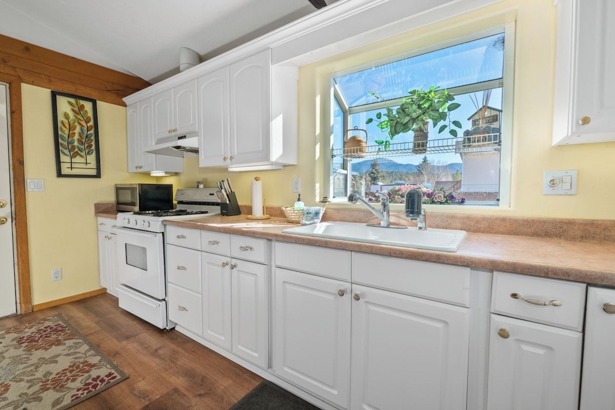 A kitchen with white cabinets, a stove, a sink, and a window with a view of greenery.