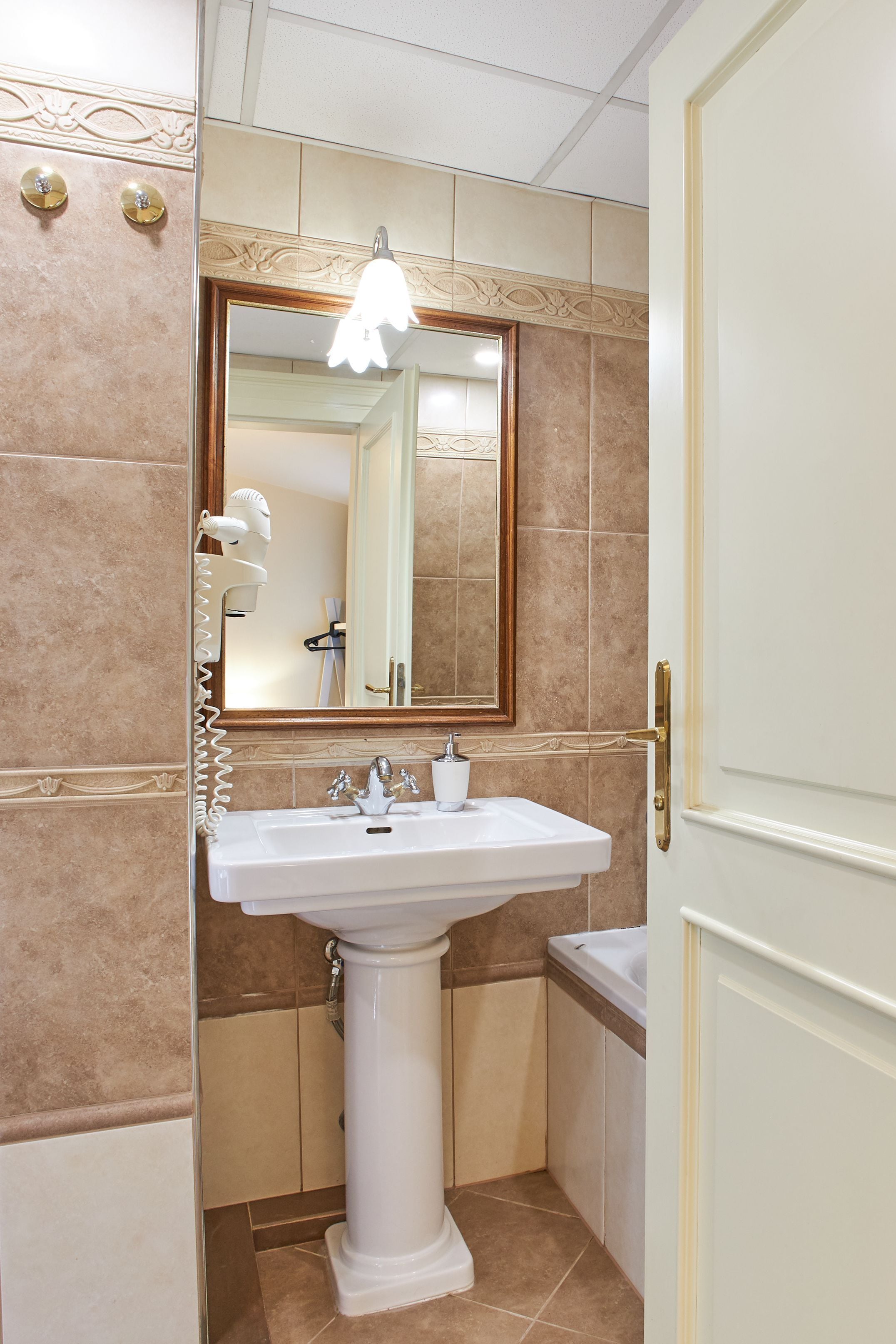 Une salle de bain avec un lavabo sur colonne, un miroir doté d’un cadre décoratif et un luminaire placé au-dessus du miroir.