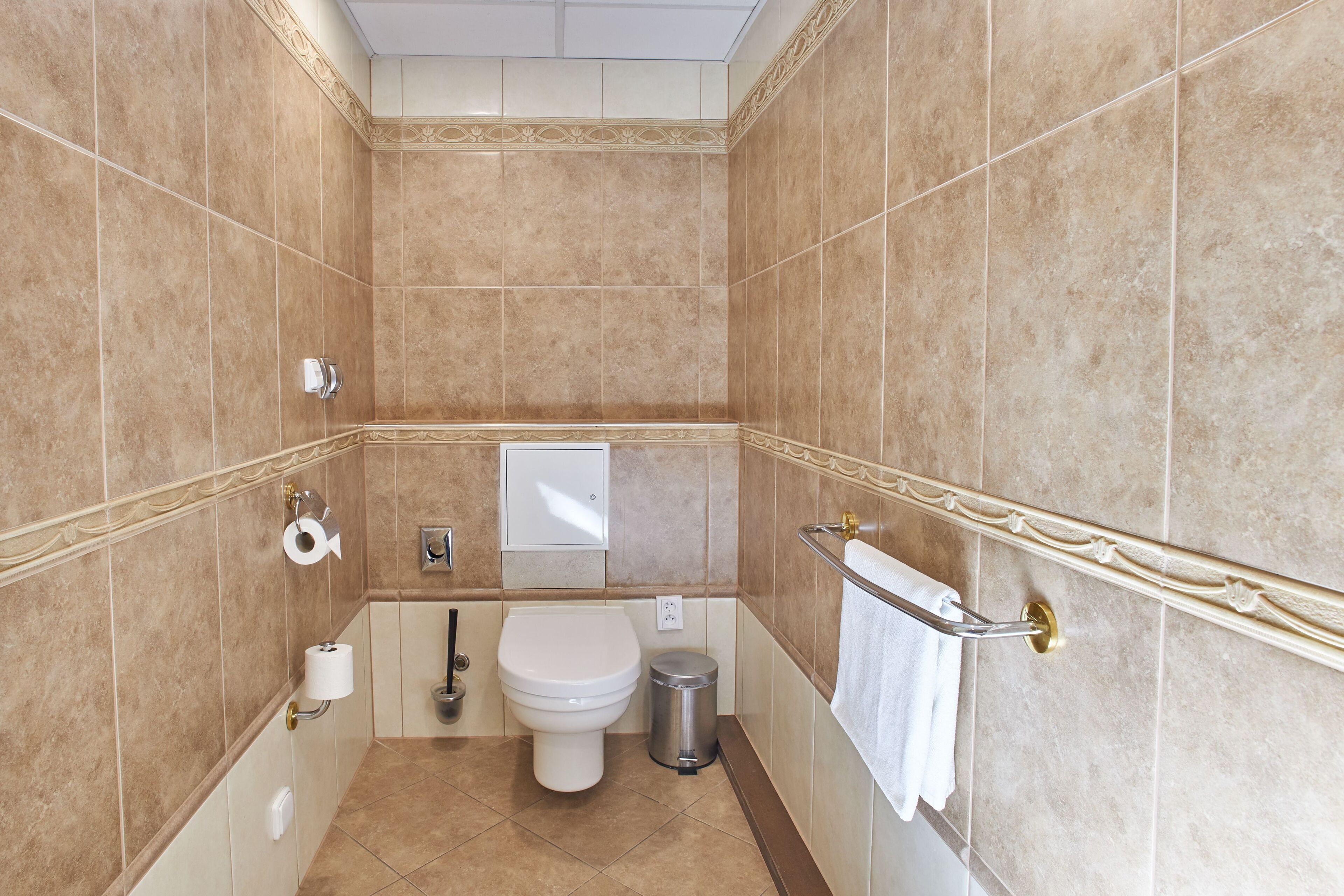 A bathroom with beige tiles, a toilet, a trash can, a towel rack, and a toilet paper holder.