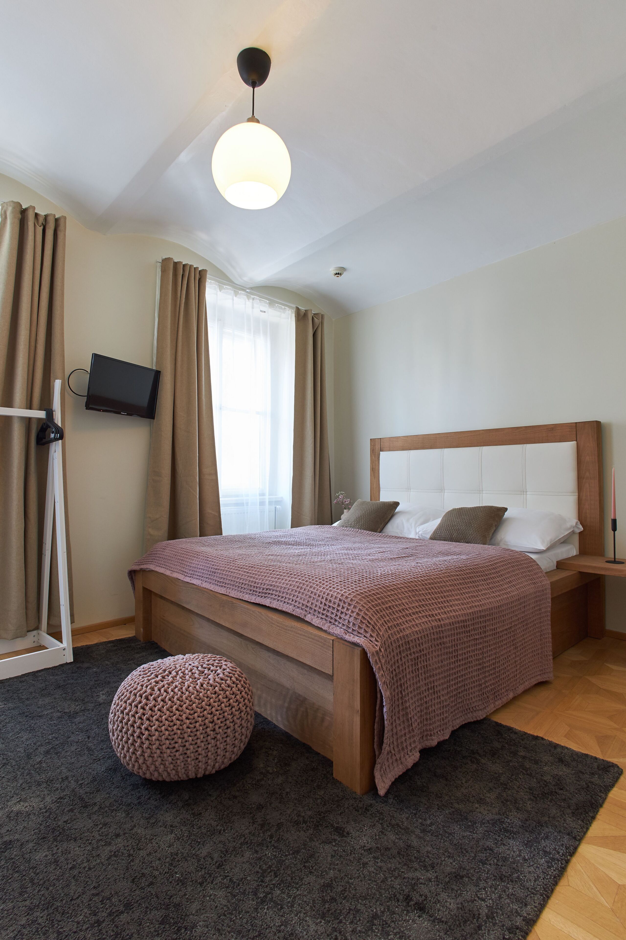 A bedroom with a wooden bed, a television mounted on the wall, and a small round ottoman.
