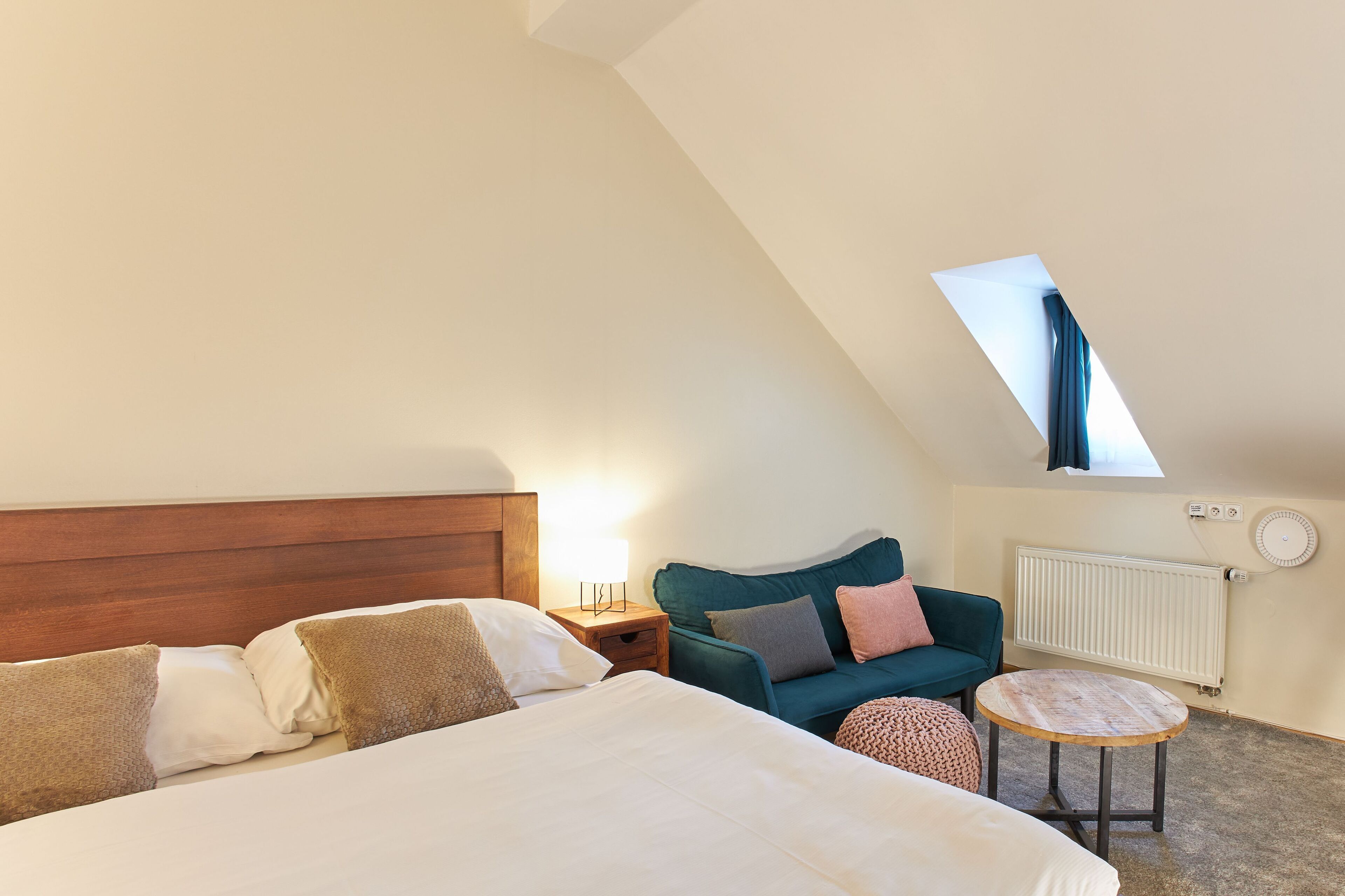 Quarto com cama grande, uma mesinha redonda, poltrona azul-esverdeada e janela com cortina.
