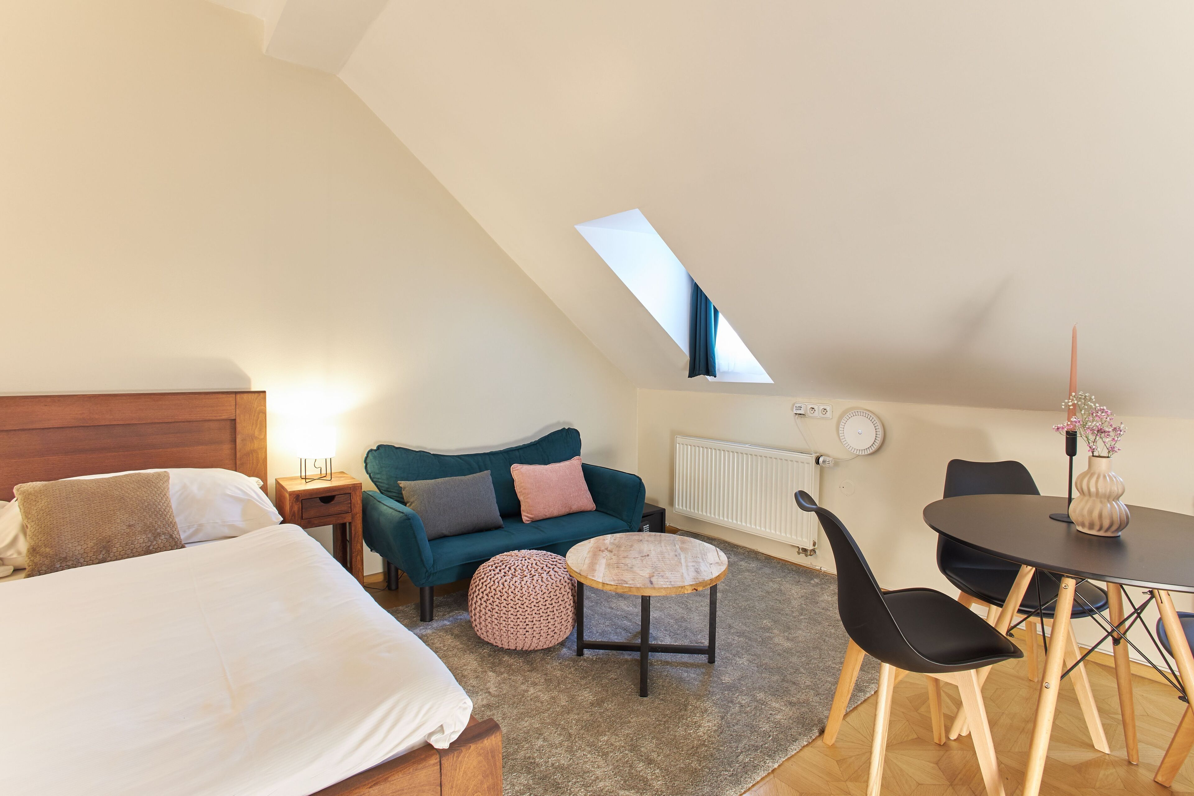 A cozy, well-lit room with a bed, a sofa, a round wooden table, and a dining table with chairs.