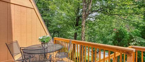 A wooden deck with a round table and chairs, surrounded by a wooden railing, overlooking a forested area.