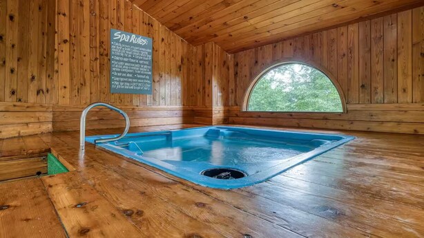 A spa room with wooden walls, a hot tub, and a window with a view of trees.