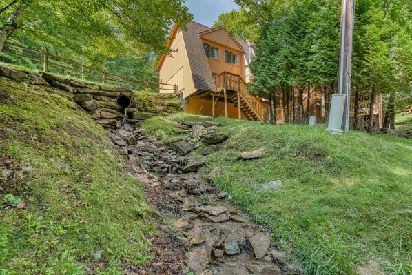Una casa con una escalera de piedra que lleva a una terraza de madera, rodeada de abundante vegetación y un muro de piedra.