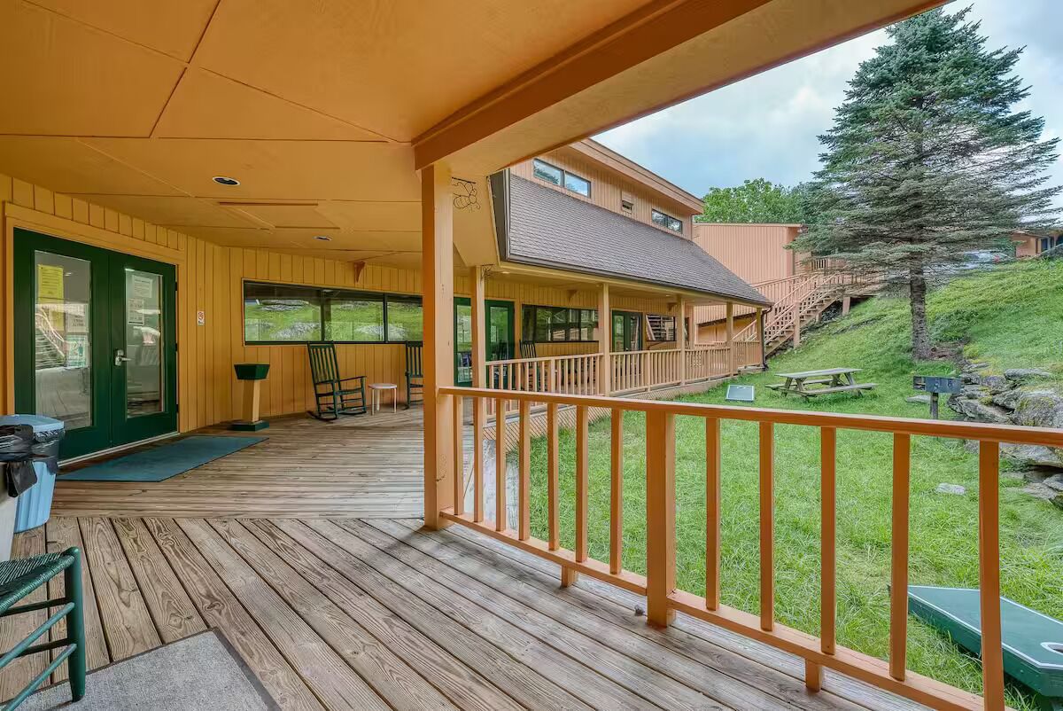 Une terrasse en bois avec une balustrade, un porche couvert, une porte verte et une vue sur une colline avec un arbre.