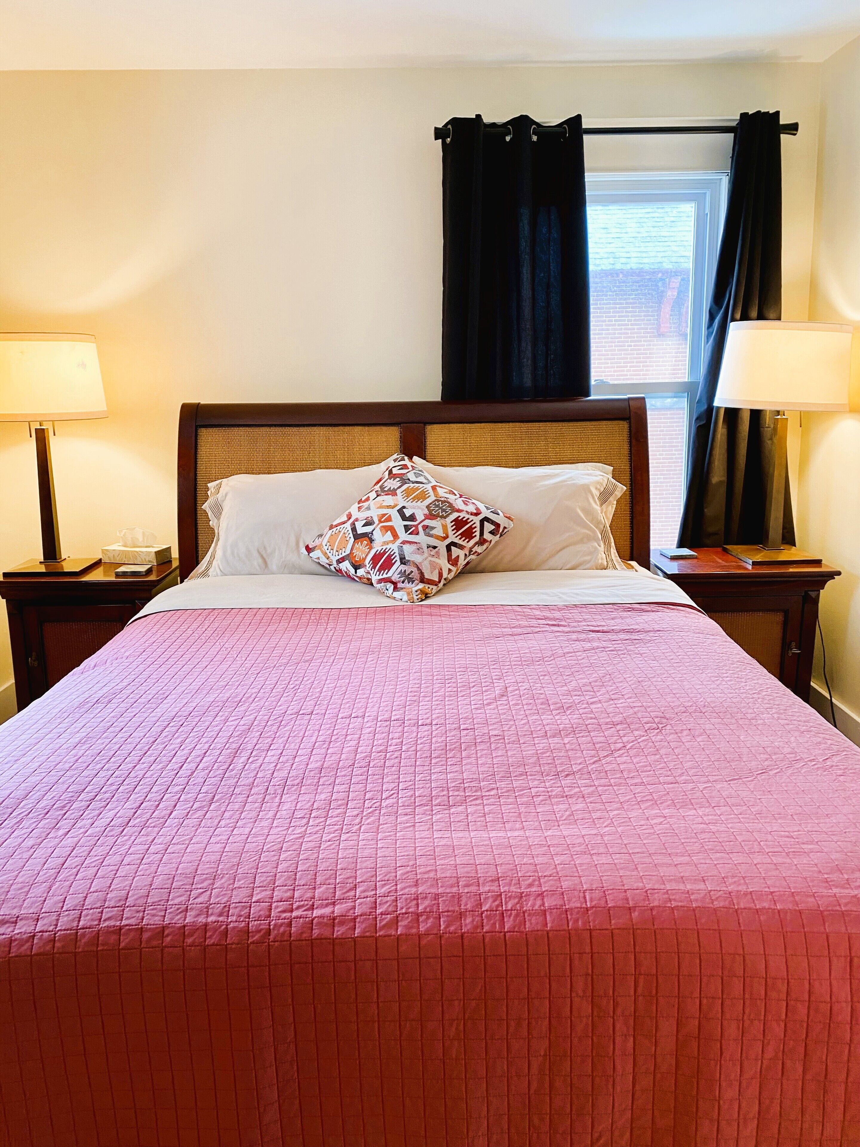 Un lit bien fait, avec une couverture rose, un coussin à motifs et deux lampes de chevet.