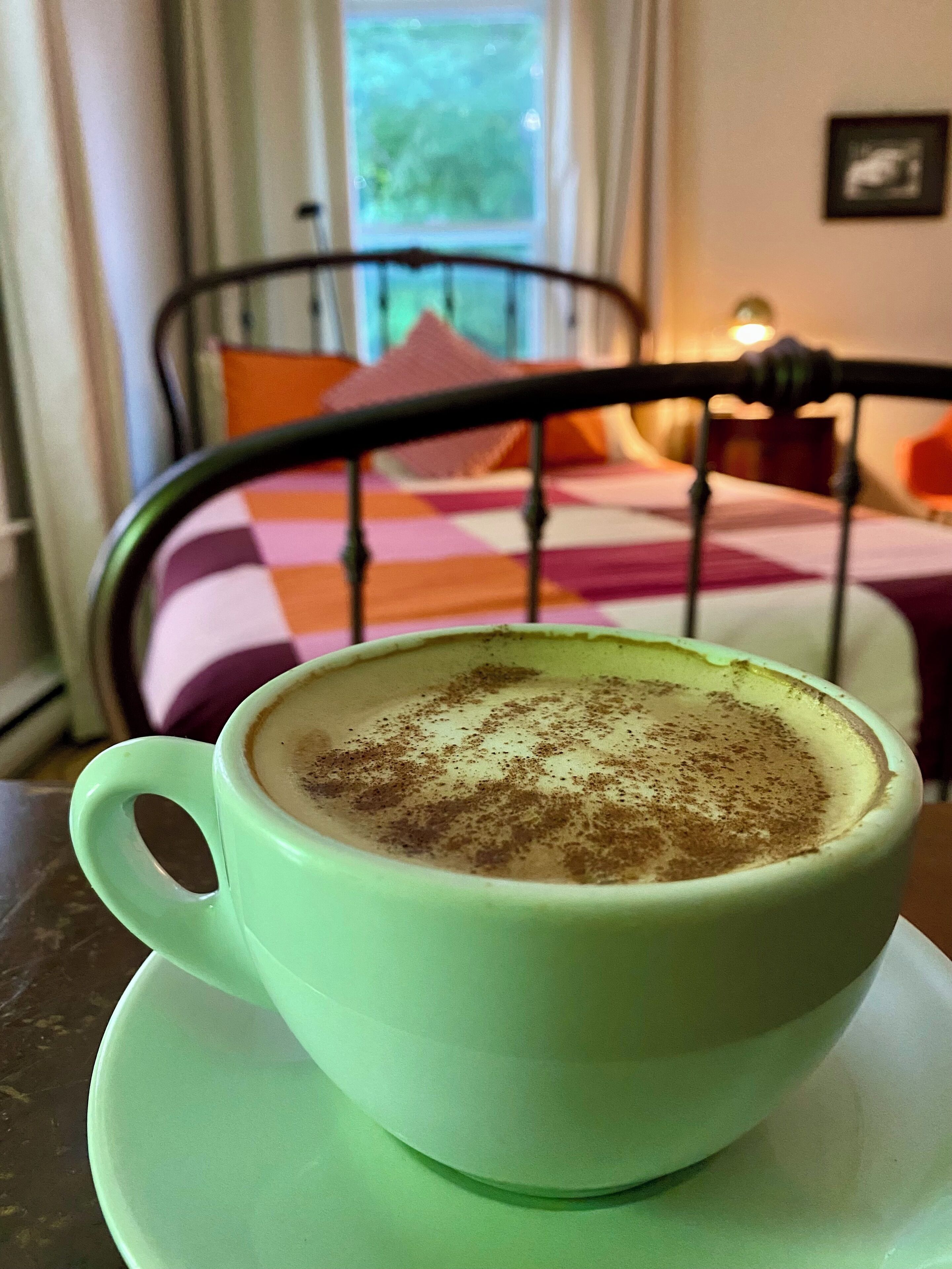 A cup of coffee on a saucer in a room with a bed and a window.