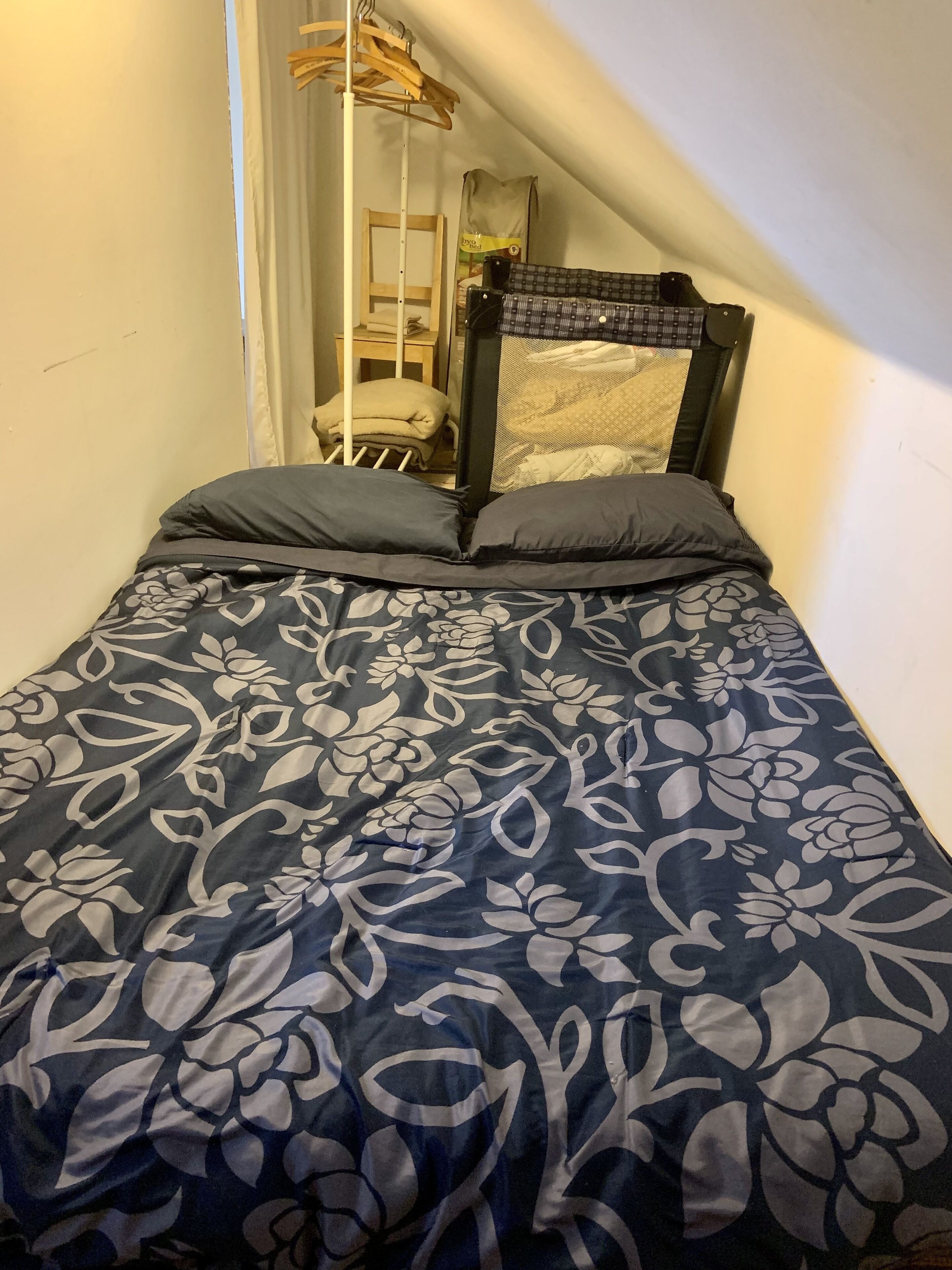 Une petite chambre avec un lit simple, recouverte d’une courtepointe à motifs floraux sombres, dotée d’une tête de lit en bois et d’une fenêtre donnant sur une chaise et un sac au fond.