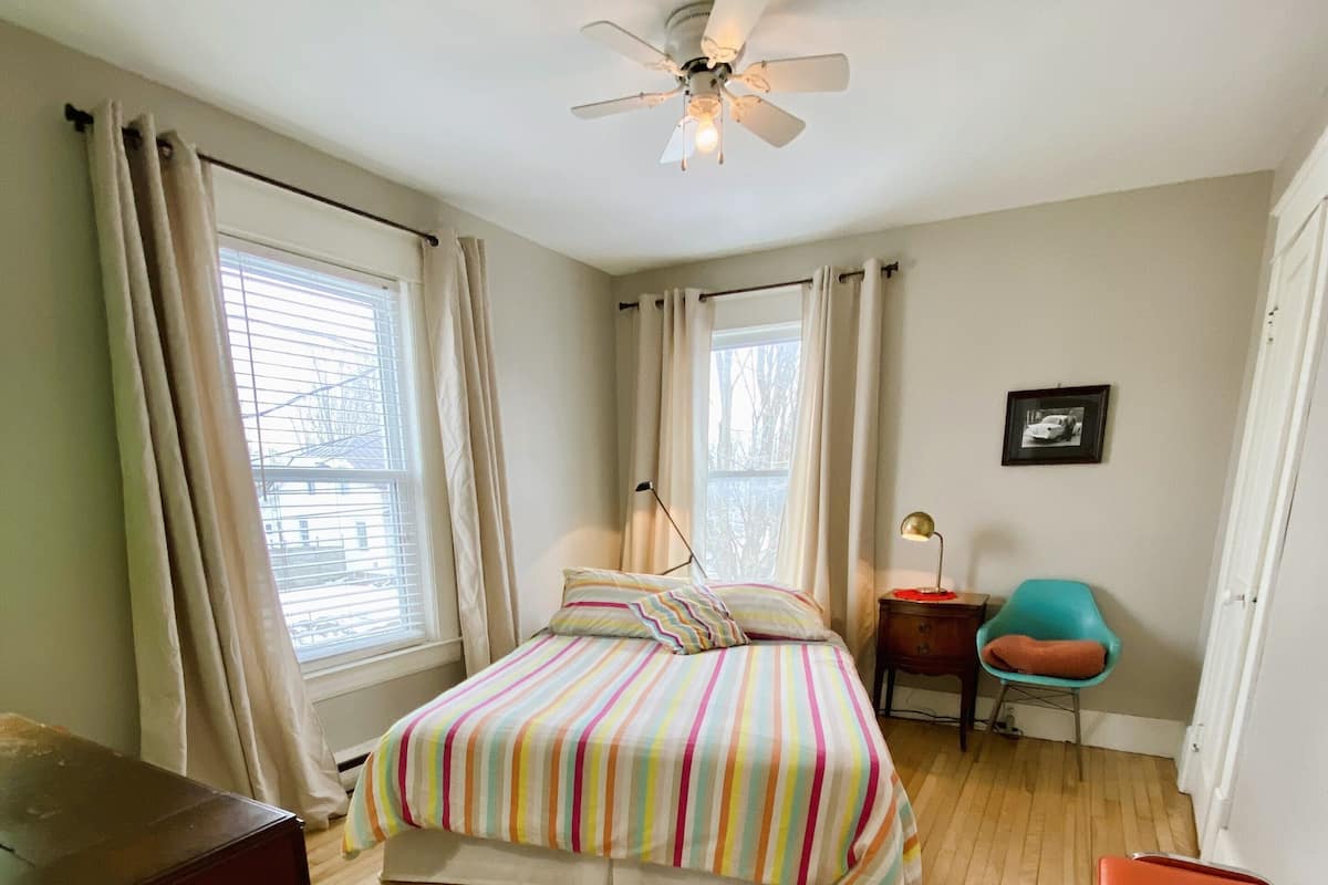 A bedroom with a bed, a desk, a chair, a ceiling fan, and a window with curtains.