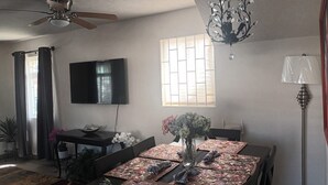A dining area with a table set for four, a chandelier, a flat-screen TV, and a window with blinds.