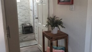 A hallway with a wooden shelf holding towels and a potted plant. A door leads to a bathroom with a shower and a towel rack.