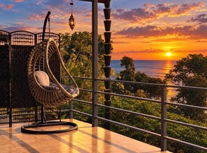 Un balcón con una silla colgante, una valla de metal y vistas al atardecer sobre el océano.