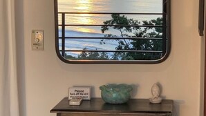 Blick durch ein Fenster auf einen Sonnenuntergang über dem Meer, ein Schild mit der Bitte, die Klimaanlage auszuschalten, und eine dekorative Vitrine mit einer Buddha-Statue.