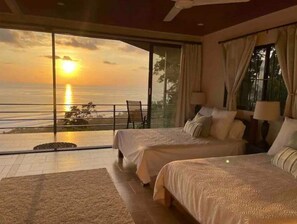A bedroom with two beds, a view of the sunset over the ocean, and a balcony with a chair.