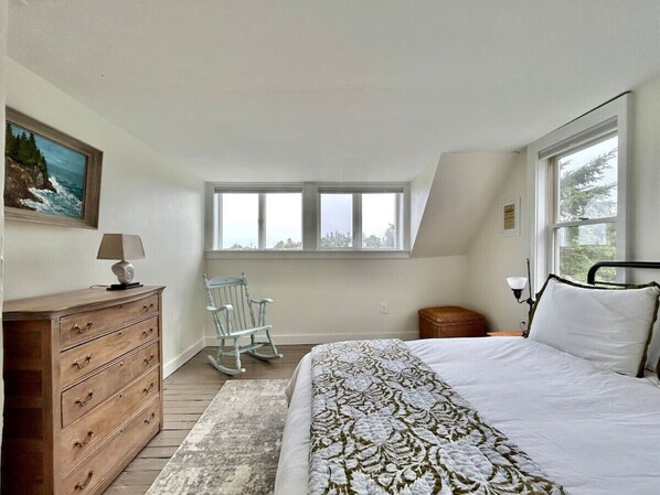 A bedroom with a bed, dresser, rocking chair, window, and a painting on the wall. - Shackford Cove Cottage (Eastport)