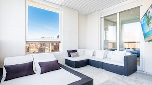 Un área de descanso moderna al aire libre con un sofá modular y dos camas individuales, ventanas amplias con vistas a la ciudad y una puerta de vidrio que da a un balcón.