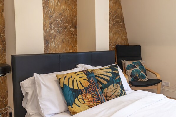 A bed with white bedding and decorative pillows, a wooden chair with a cushion, and a wall with vertical stripes.