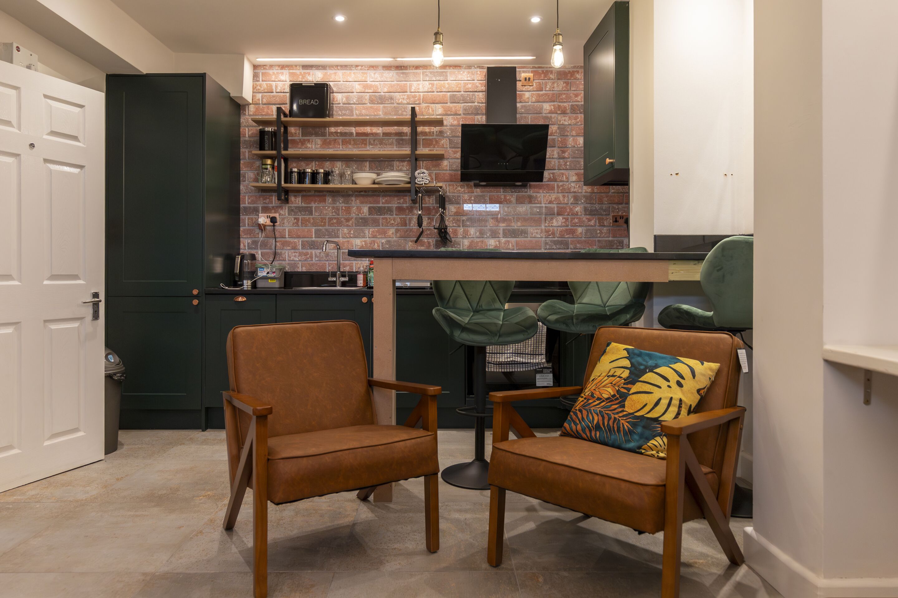 A compact kitchen with a brick backsplash, a flat-screen TV, and a dining area with two chairs and a table.