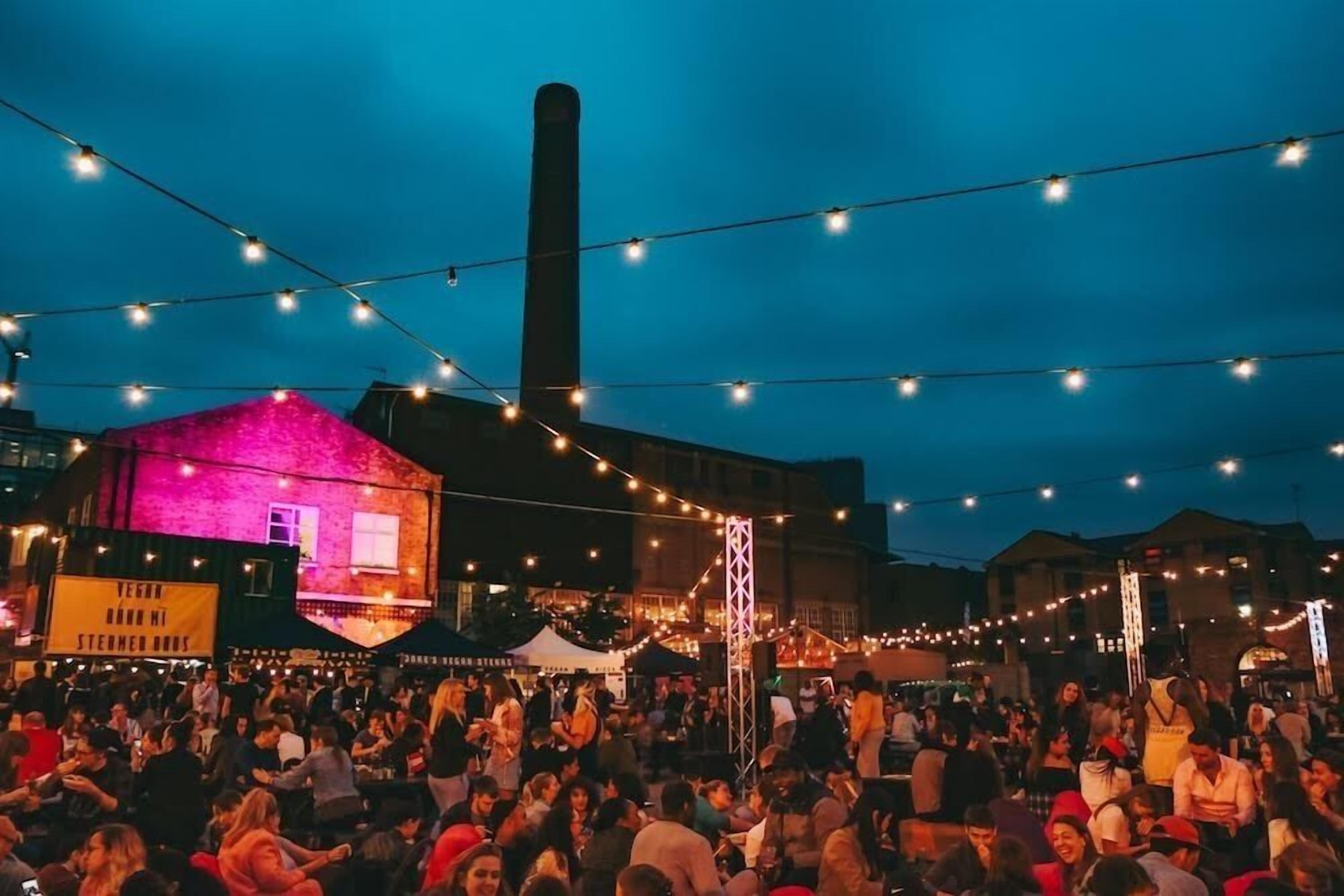 A crowded outdoor event with string lights and a building with a pink-lit facade.