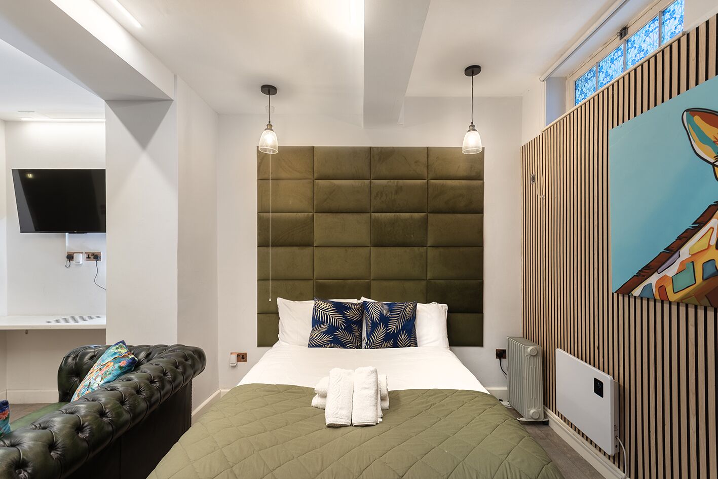 A modern hotel room with a green quilted bed, a leather sofa, a flat-screen TV, and a painting on the wall.
